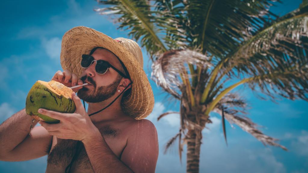 Photo of a shirtless man in a hat, drinking from a coconut with a straw